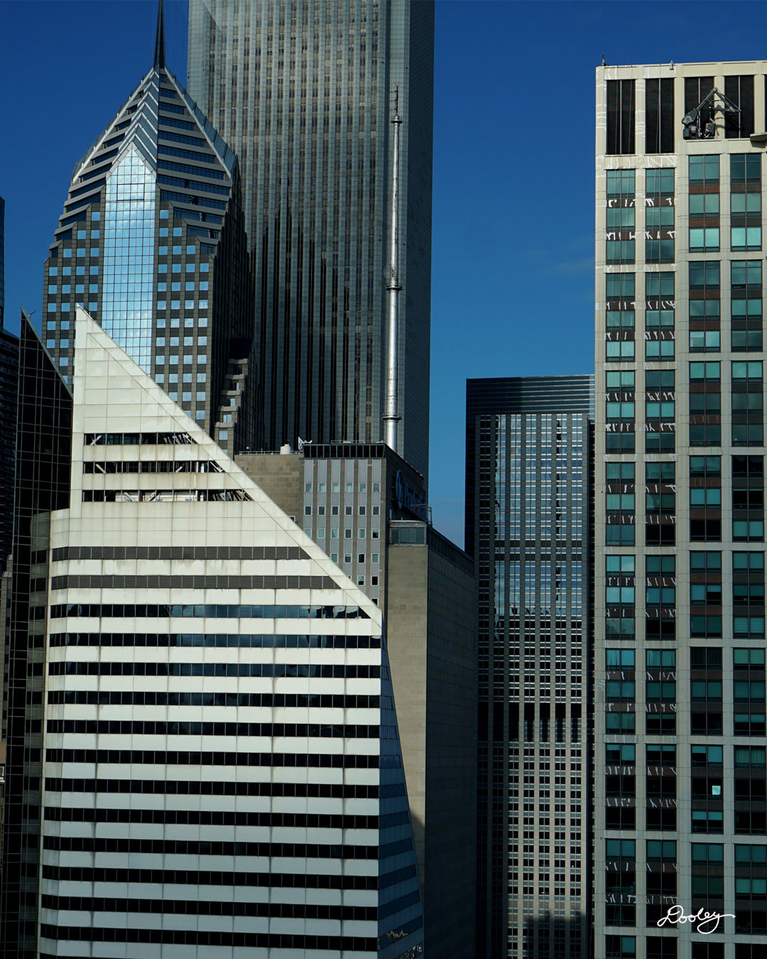 Image showcasing a cluster of towering skyscrapers in Chicago, with a vibrant blue sky as a backdrop. The buildings exhibit a variety of architectural styles, with sleek glass facades and modern designs dominating the skyline. A particularly striking feature is a diamond-shaped skyscraper, Crain Communications Building, with its unique silhouette standing out amidst the other buildings. Buildings also include Two Prudential Plaza, and a host of smaller buildings.