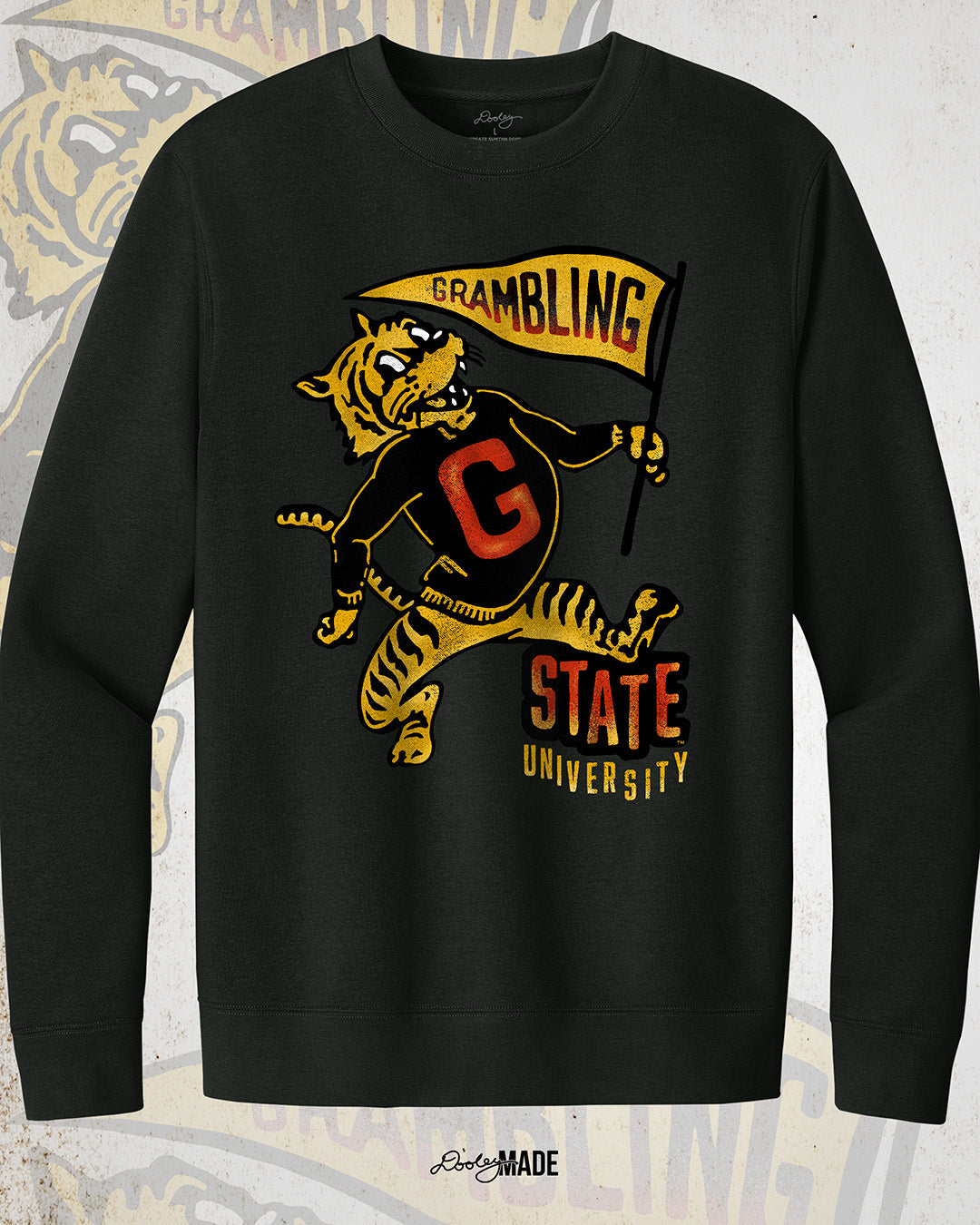 This is an image of a black crewneck sweatshirt featuring a retro-style design celebrating Grambling State University. The design includes a vintage illustration of a tiger mascot holding a yellow pennant that reads "Grambling" in bold letters. The tiger wears a shirt with a large red "G." Under the tiger is the text "State University" in red and yellow, giving it a distressed, classic look. The background features a faint overlay of the same tiger mascot design for added texture with the Dooley Made logo.