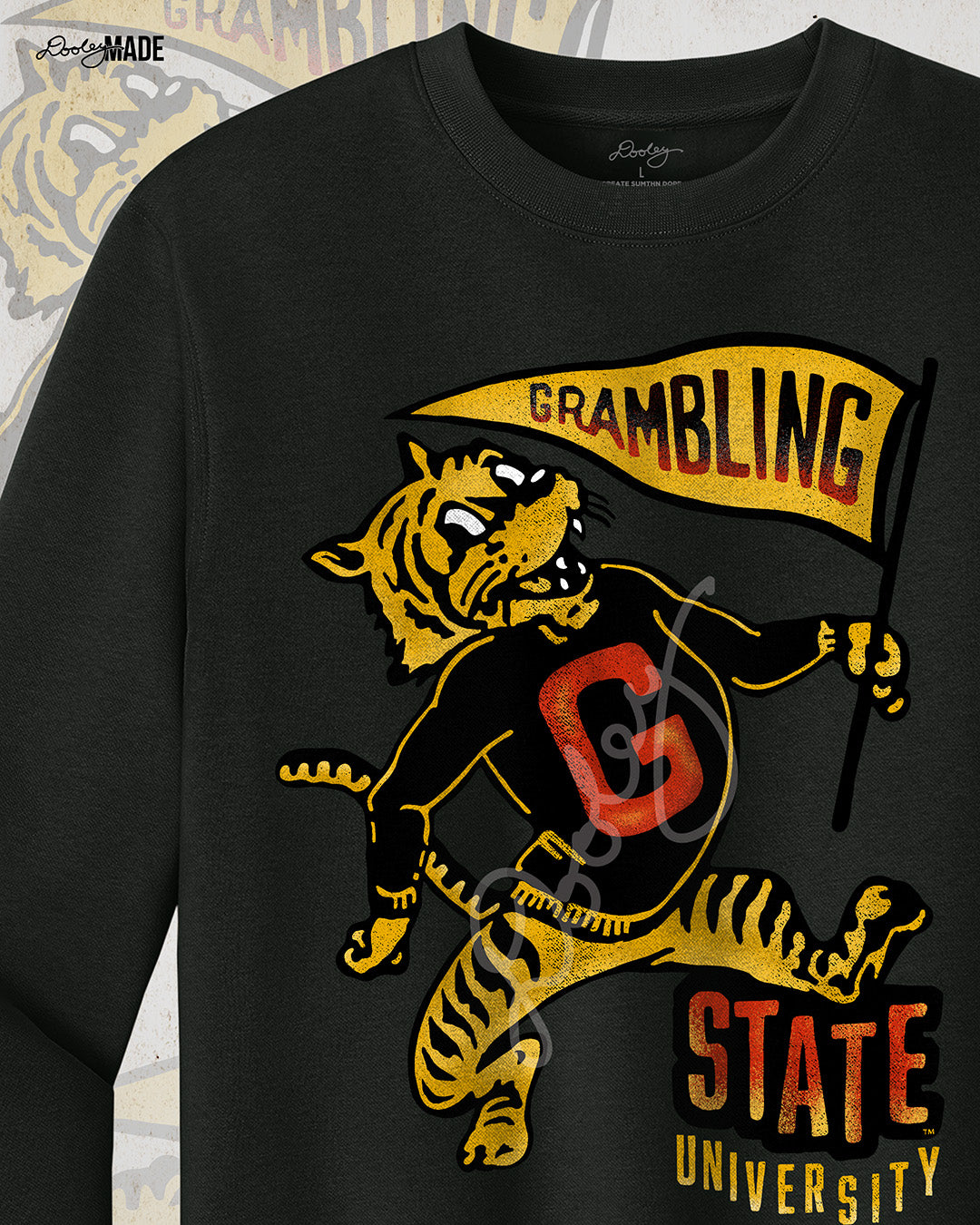 This is an image of a black crewneck sweatshirt featuring a retro-style design celebrating Grambling State University. The design includes a vintage illustration of a tiger mascot holding a yellow pennant that reads "Grambling" in bold letters. The tiger wears a shirt with a large red "G." Under the tiger is the text "State University" in red and yellow, giving it a distressed, classic look. The background features a faint overlay of the same tiger mascot design for added texture with the Dooley Made logo.