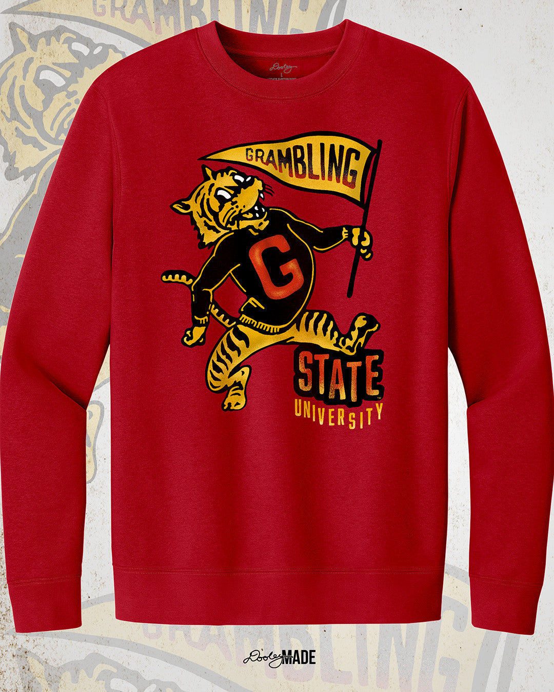 This is an image of a red crewneck sweatshirt featuring a retro-style design celebrating Grambling State University. The design includes a vintage illustration of a tiger mascot holding a yellow pennant that reads "Grambling" in bold letters. The tiger wears a shirt with a large red "G." Under the tiger is the text "State University" in red and yellow, giving it a distressed, classic look. The background features a faint overlay of the same tiger mascot design for added texture with the Dooley Made logo.