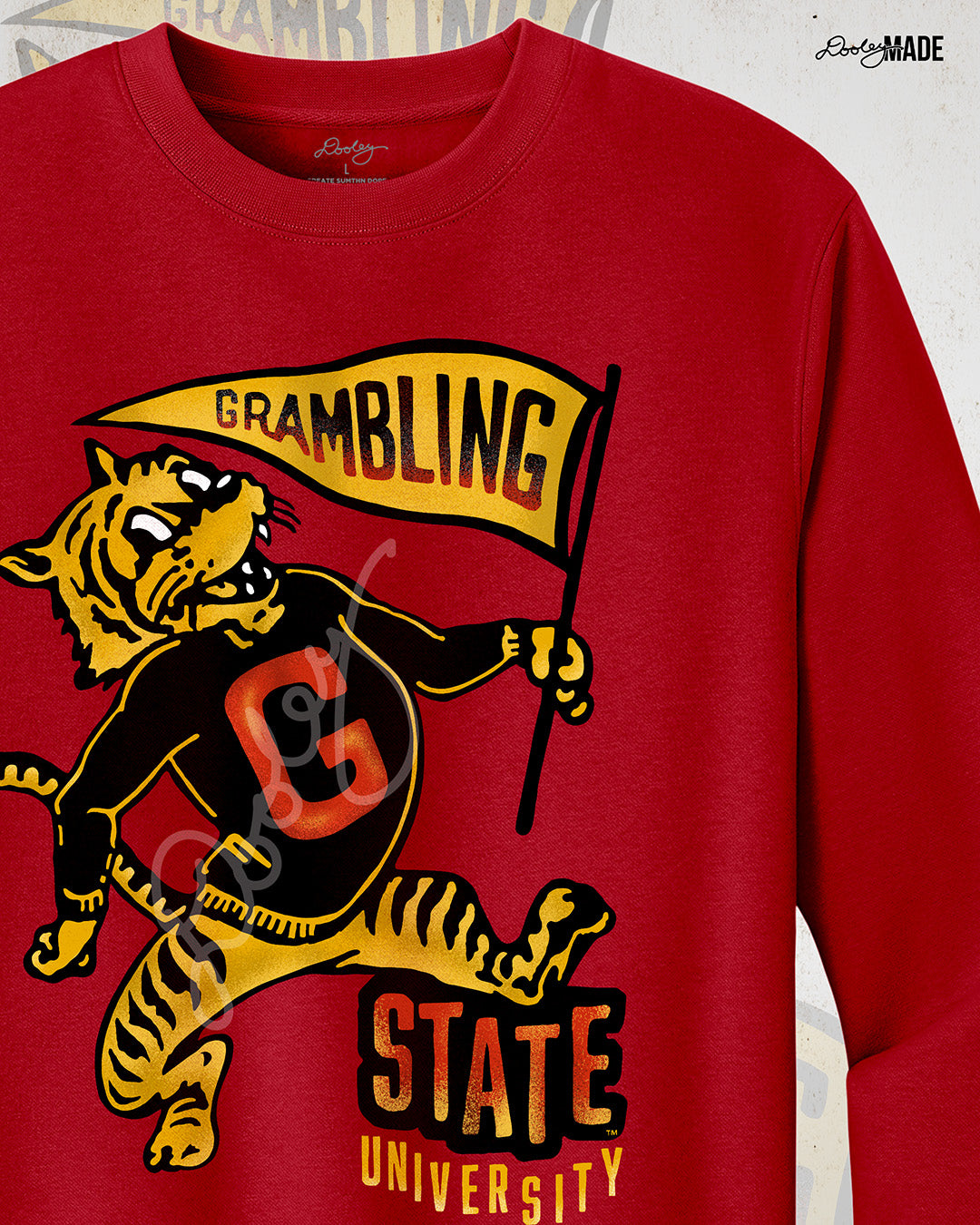 This is an image of a red crewneck sweatshirt featuring a retro-style design celebrating Grambling State University. The design includes a vintage illustration of a tiger mascot holding a yellow pennant that reads "Grambling" in bold letters. The tiger wears a shirt with a large red "G." Under the tiger is the text "State University" in red and yellow, giving it a distressed, classic look. The background features a faint overlay of the same tiger mascot design for added texture with the Dooley Made logo.