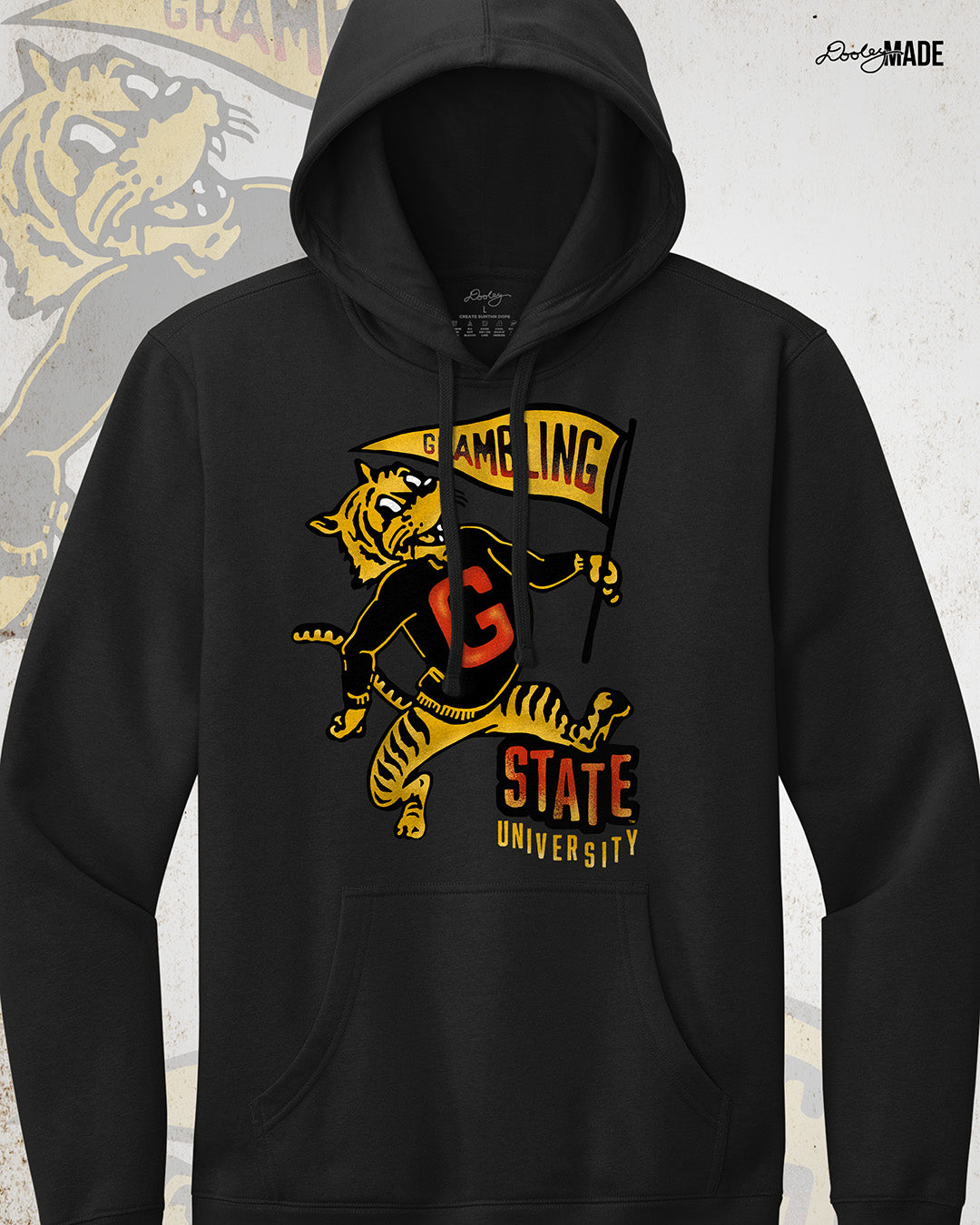 This is an image of a premium black hoodie featuring a retro-style design celebrating Grambling State University. The design includes a vintage illustration of a tiger mascot holding a yellow pennant that reads "Grambling" in bold letters. The tiger wears a shirt with a large red "G." Under the tiger is the text "State University" in red and yellow, giving it a distressed, classic look. The background features a faint overlay of the same tiger mascot design for added texture with the Dooley Made logo.