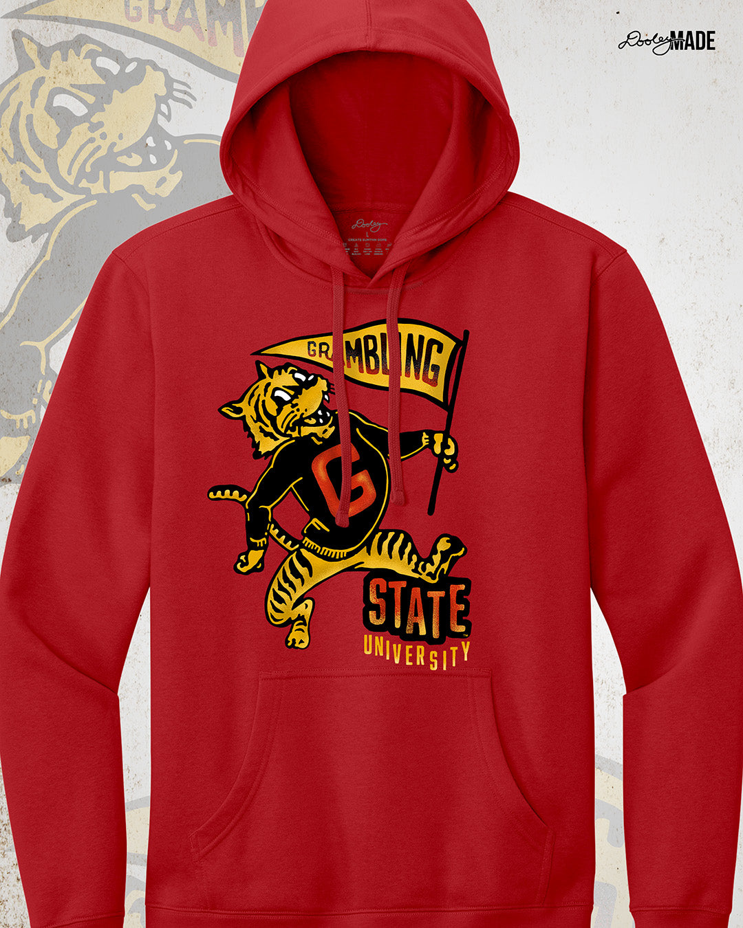 This is an image of a premium red hoodie featuring a retro-style design celebrating Grambling State University. The design includes a vintage illustration of a tiger mascot holding a yellow pennant that reads "Grambling" in bold letters. The tiger wears a shirt with a large red "G." Under the tiger is the text "State University" in red and yellow, giving it a distressed, classic look. The background features a faint overlay of the same tiger mascot design for added texture with the Dooley Made logo.