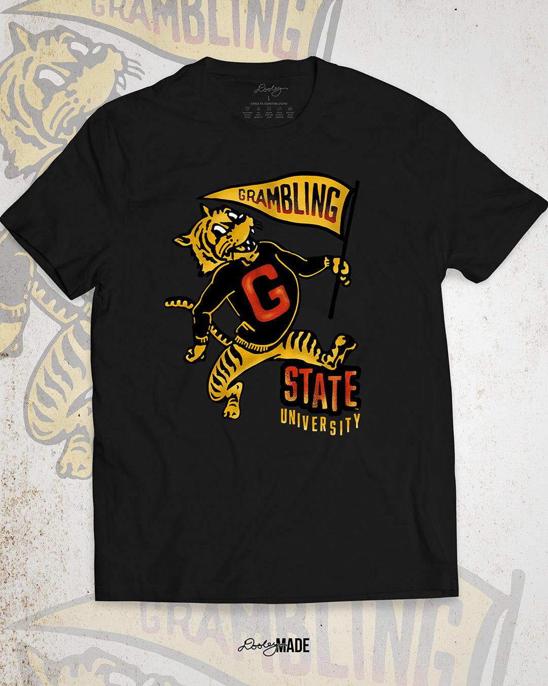 This is an image of a premium black shirt featuring a retro-style design celebrating Grambling State University. The design includes a vintage illustration of a tiger mascot holding a yellow pennant that reads "Grambling" in bold letters. The tiger wears a shirt with a large red "G." Under the tiger is the text "State University" in red and yellow, giving it a distressed, classic look. The background features a faint overlay of the same tiger mascot design for added texture with the Dooley Made logo.