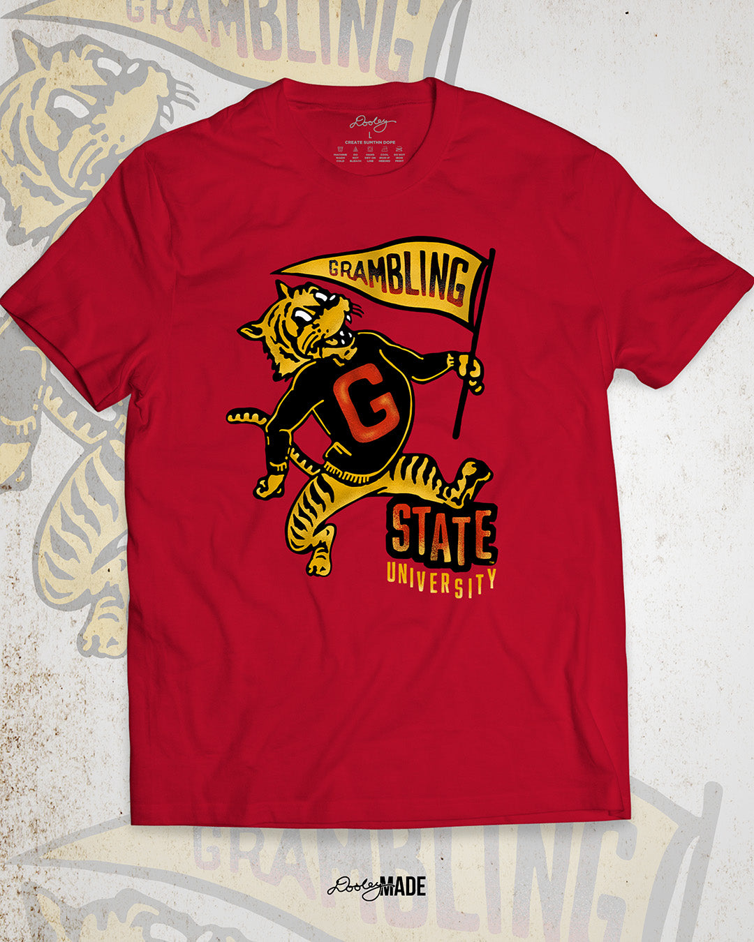 This is an image of a premium red shirt featuring a retro-style design celebrating Grambling State University. The design includes a vintage illustration of a tiger mascot holding a yellow pennant that reads "Grambling" in bold letters. The tiger wears a shirt with a large red "G." Under the tiger is the text "State University" in red and yellow, giving it a distressed, classic look. The background features a faint overlay of the same tiger mascot design for added texture with the Dooley Made logo.