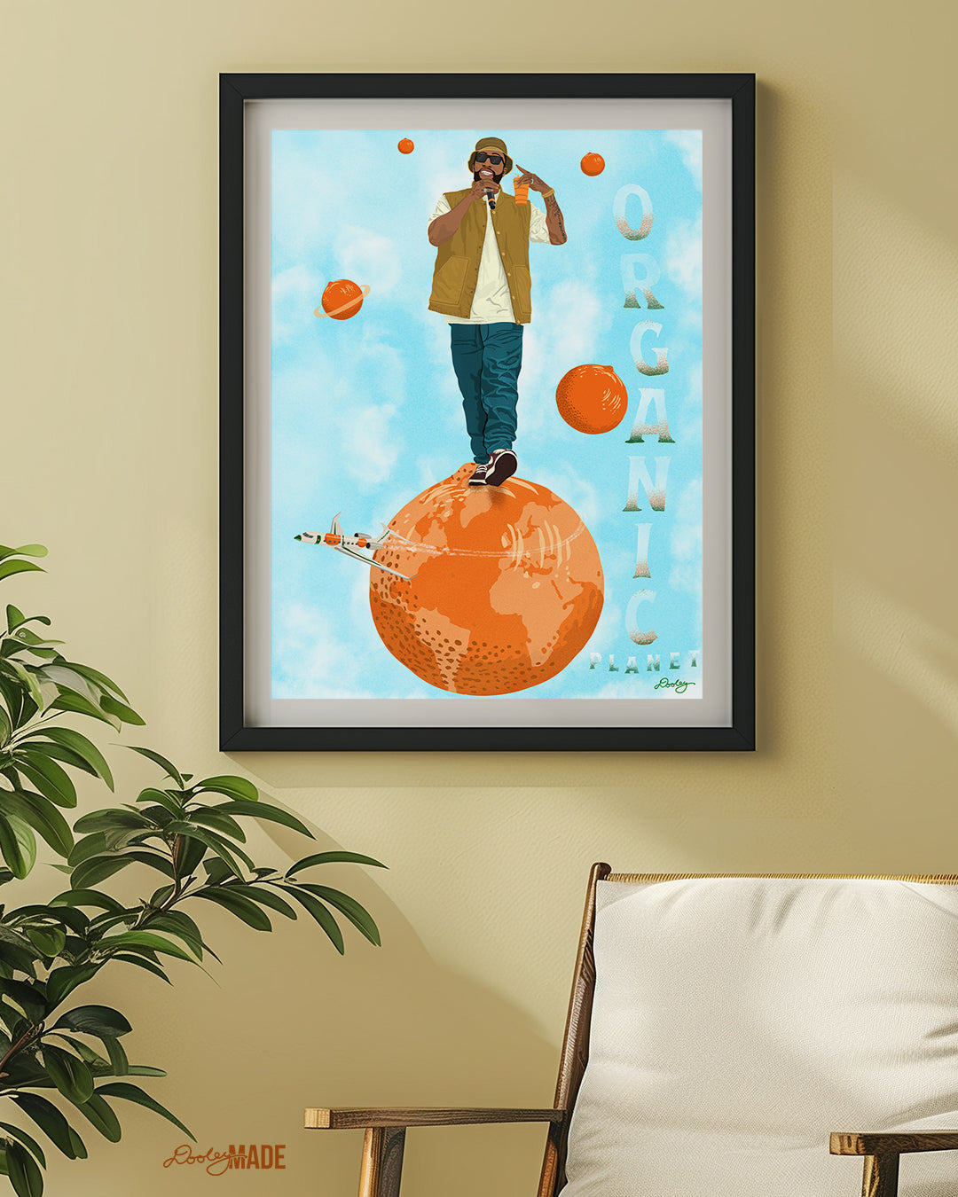 An image of a Larry June illustration inside of a black frame on a tan-yellow wall above a plant and sitting chair. The image shows him standing on an orange planet with floating orange planets, alongside the text "Organic Planet." There is also a orange themed jet flying from the right to left on the image.