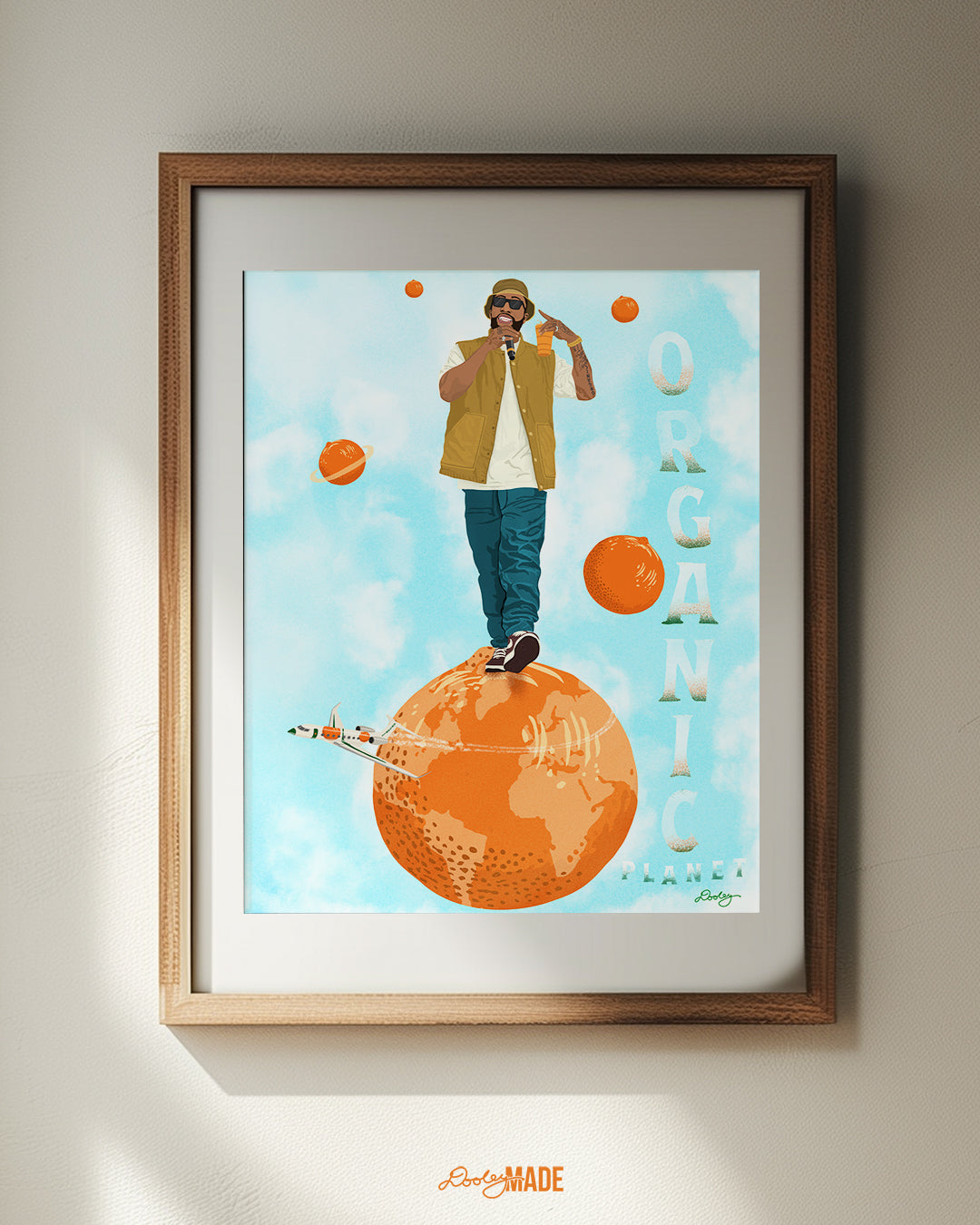 An image of a Larry June illustration inside of a wooden frame a white wall. The image shows him standing on an orange planet with floating orange planets, alongside the text "Organic Planet." There is also a orange themed jet flying from the right to left on the image.