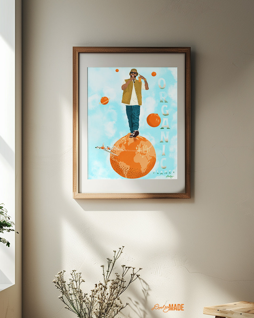 An image of a Larry June illustration inside of a wooden frame a white wall. The image shows him standing on an orange planet with floating orange planets, alongside the text "Organic Planet." There is also a orange themed jet flying from the right to left on the image.