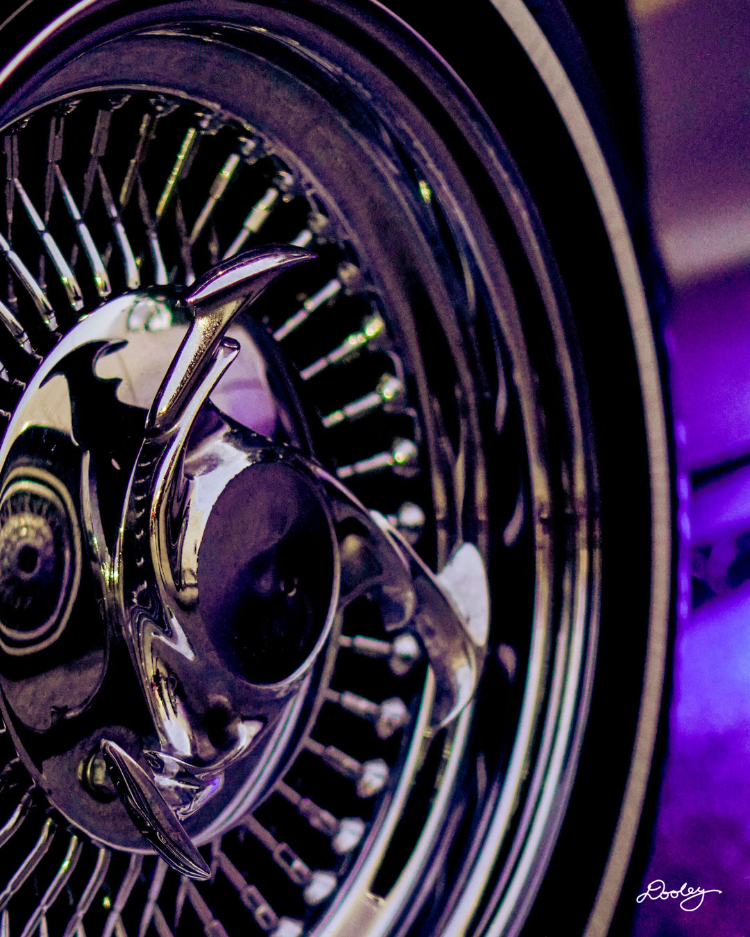 The image shows a close-up of a custom wire wheel, specifically a set of Dayton wire wheels. The wheel is chrome-plated and has a large number of spokes radiating out from the center. The center cap is also chrome-plated and features a distinctive design with two curved horns. The wheel is mounted on a white-walled tire and appears to be illuminated by purple light. Photo taken by Dre Dooley