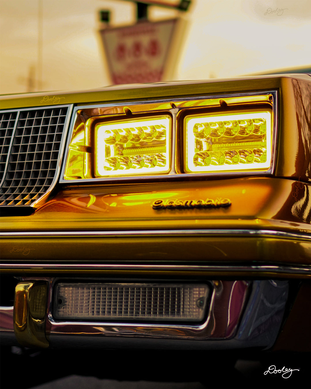 The image shows the front of a classic 1980s Oldsmobile. The car has a dark gold paint job and features distinctive rectangular headlights with a bright yellow hue. The Oldsmobile emblem is visible on the front grille. There is a  faded orange-tan sky and a sign behind the car.