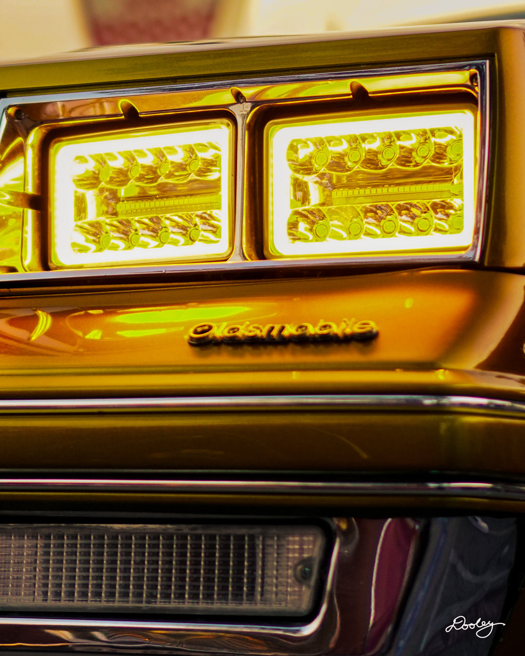 The image shows the front of a classic 1980s Oldsmobile. The car has a bright gold paint job and features distinctive rectangular headlights with a bright yellow hue. The Oldsmobile emblem is visible on the front grille.