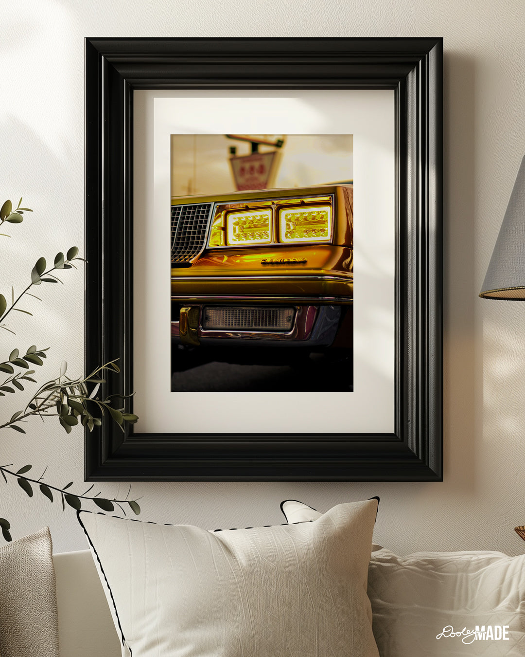 The image shows a close-up of a classic 1980s Oldsmobile's car's front end. The car is a beautiful golden color, and the headlights are on, casting a warm glow. The image is framed and hanging on a wall in a modern living room setting.