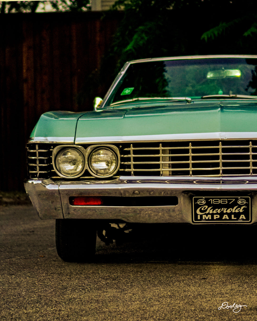 The image shows a classic 1967 Chevrolet Impala in a beautiful shade of green. The car is parked on dark pavement, with a dark background that highlights its sleek lines and chrome accents. The Impala's distinctive features, such as the double headlights, iconic grille, and "Chevrolet" emblem, are clearly visible. The car looks well-maintained and shines beautifully in the available light.