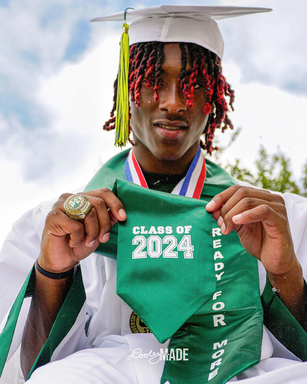 A photo advertising Personalized Graduation Photography showing a graduate wearing a white cap and gown and green stole against a beautiful cloudy blue and white sky. The graduate is holding the stole showing the words "class of 2024."