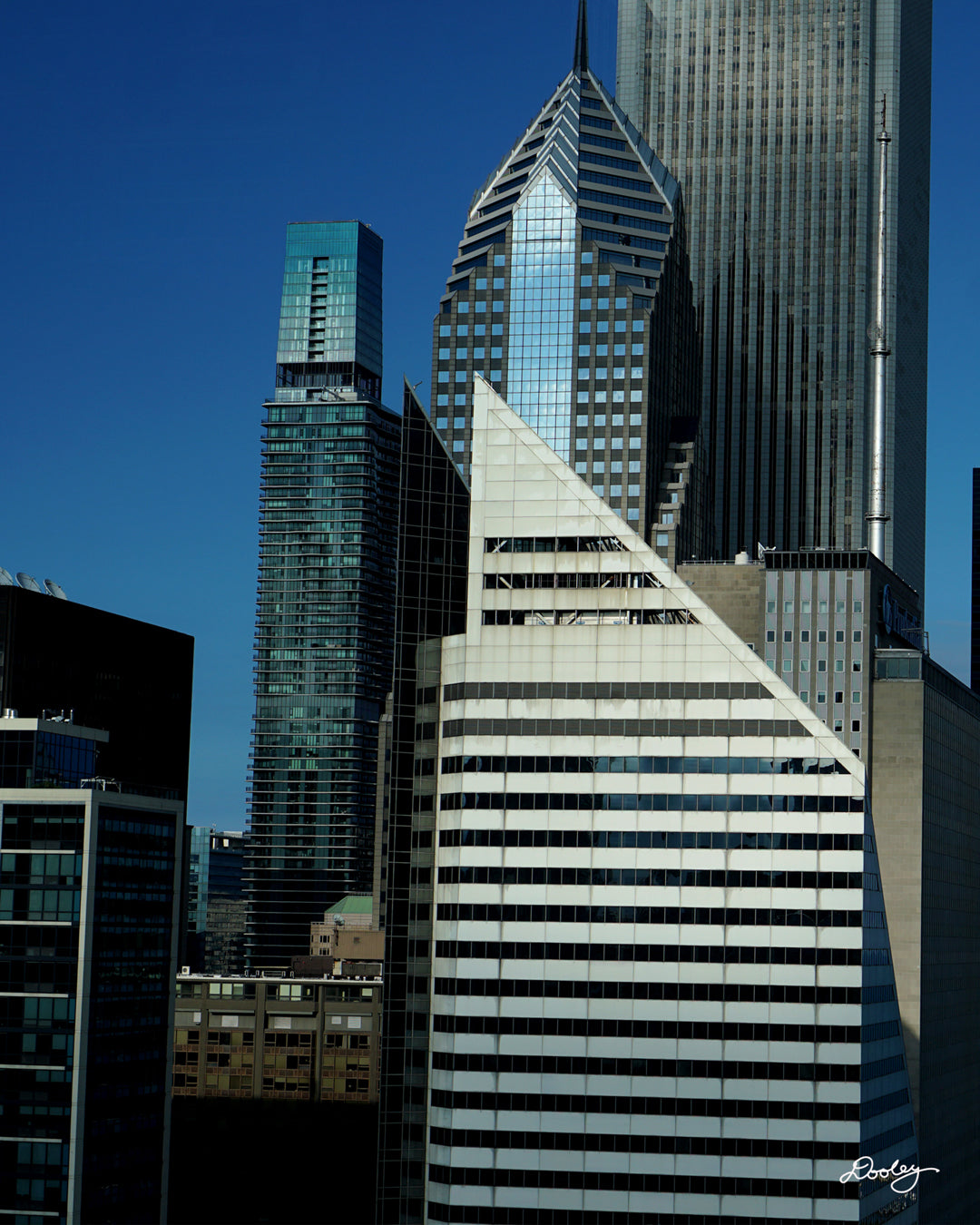 Image showcasing a cluster of towering skyscrapers in Chicago, with a vibrant blue sky as a backdrop. The buildings exhibit a variety of architectural styles, with sleek glass facades and modern designs dominating the skyline. A particularly striking feature is a diamond-shaped skyscraper, Crain Communications Building, with its unique silhouette standing out amidst the other buildings. Buildings also include Two Prudential Plaza, The St. Regis Chicago, and a host of smaller buildings.