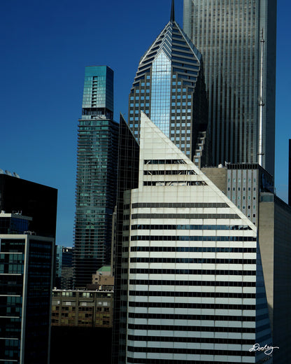 Image showcasing a cluster of towering skyscrapers in Chicago, with a vibrant blue sky as a backdrop. The buildings exhibit a variety of architectural styles, with sleek glass facades and modern designs dominating the skyline. A particularly striking feature is a diamond-shaped skyscraper, Crain Communications Building, with its unique silhouette standing out amidst the other buildings. Buildings also include Two Prudential Plaza, The St. Regis Chicago, and a host of smaller buildings.