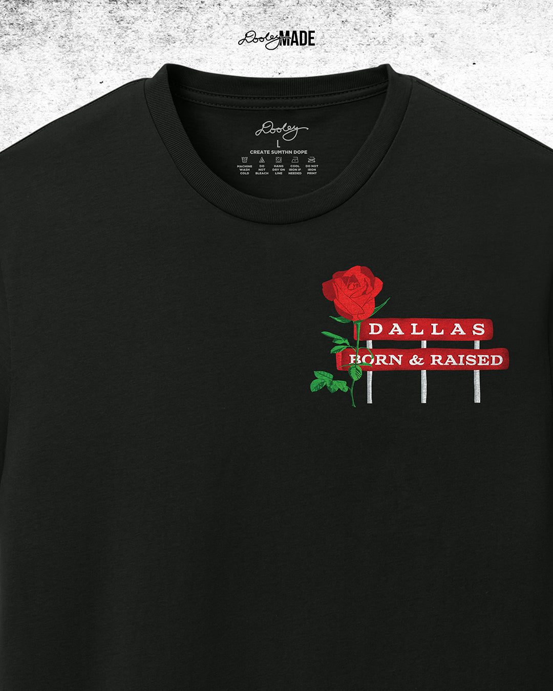 An image of premium black shirt from The Dallas Concrete Rose Collection featuring a vibrant illustration by Dre Dooley of a Rose and a sign reading "Dallas Born and Raised" on the left chest. Zoomed to show details.