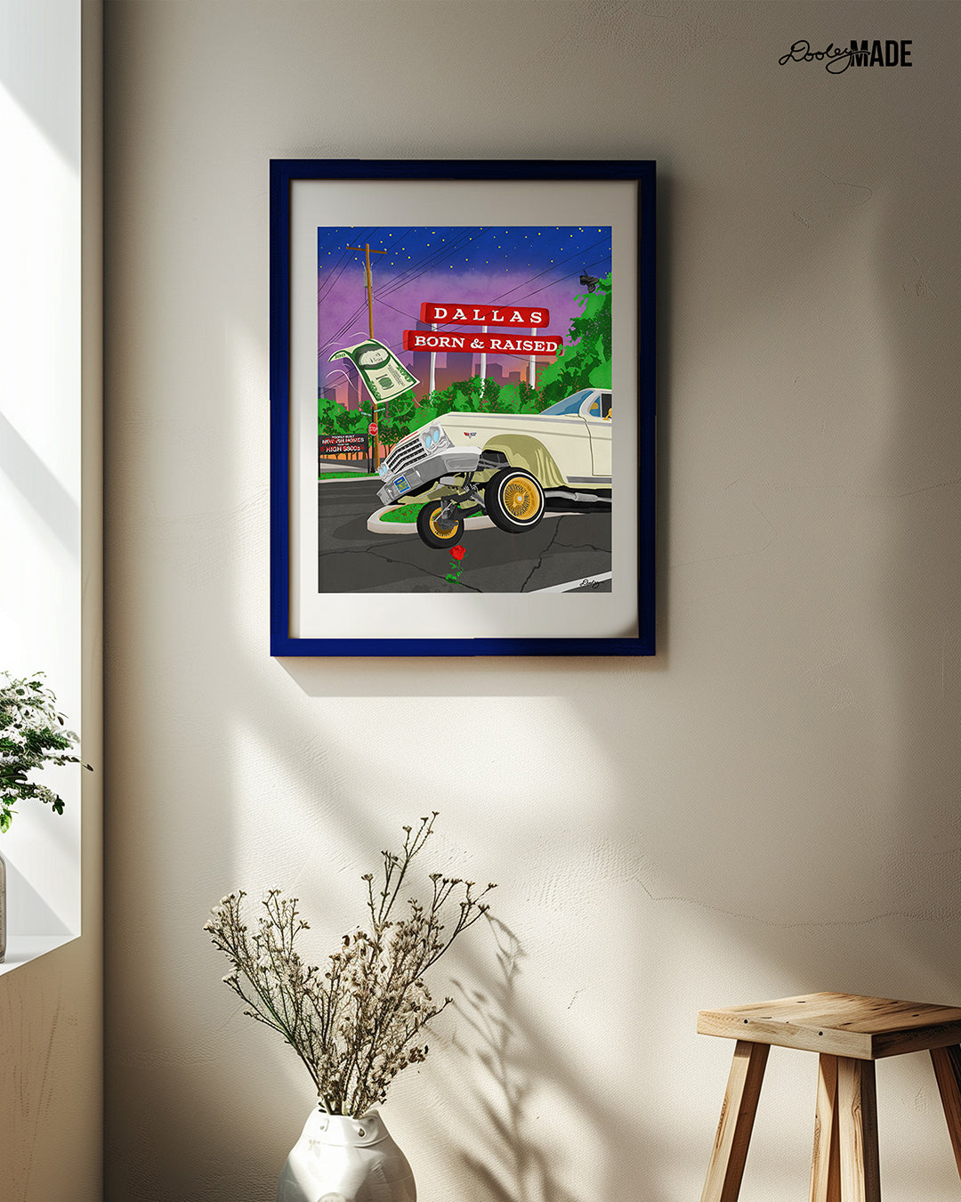 A frame on a wall holding a print of The Dallas Concrete Rose artwork - a vibrant illustration by Dre Dooley of a classic 1964 Chevy Impala lowrider with gold wire wheels, hopping in the streets of Dallas. A red street sign reads "Dallas Born & Raised", set against a sunset city skyline. A floating $100 bill, power lines, and urban elements reflect the culture of Dallas, TX. A single red rose grows from a cracked street, symbolizing resilience and roots.