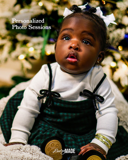 A brown skinned baby in a green checkered outfit with a white bow, surrounded by festive holiday decorations, advertising Personalized Photo Sessions in the Dallas DFW Metroplex by Dooley Made.