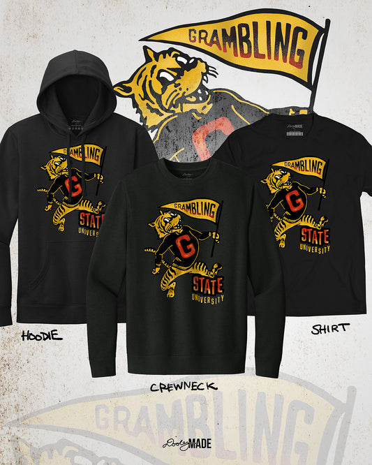 This is an image of a premium black hoodie, crewneck sweater, and shirt featuring a retro-style design celebrating Grambling State University. The design includes a vintage illustration of a tiger mascot holding a yellow pennant that reads "Grambling" in bold letters. The tiger wears a shirt with a large red "G." Under the tiger is the text "State University" in red and yellow, giving it a distressed, classic look. The background features a faint overlay of the same tiger mascot design for added texture. 