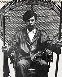 An zoomed image of Huey P. Newton artwork showing him sitting in a woven chair wearing a black leather jacket, dark colored pants, and a black cap while holding the barrel of a rifle in one hand and a wooden spear in the other.