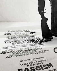 A zoomed image of Huey P. Newton artwork featuring a poster advertising the Black Panther Party's Free Breakfast Program, Liberation School, Community Politics Education Classes stretched in perspective across the floor under the butt of a gun and group of shotgun shells. It also shows words "National Conference for a United front against Fascism. 