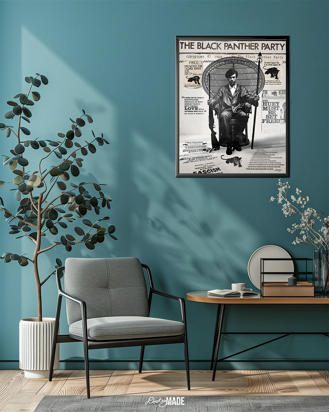 An image of framed Huey P. Newton artwork featuring the Black Panther Party's 10 Point Program, a poster that hung inside the door of the Chicago Chapter headquarters, the quote "I believe what motivates people is not great hate, but great love for other people," and other historical memorabilia. The frame is on the blue-teal wall in a living room setting above a small table chair, and tree plant.