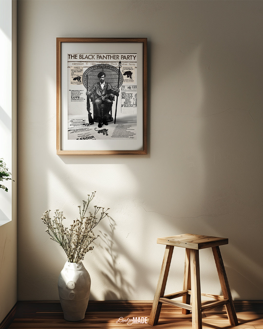 An image of framed Huey P. Newton artwork featuring the Black Panther Party's 10 Point Program, a poster that hung inside the door of the Chicago Chapter headquarters, the quote "I believe what motivates people is not great hate, but great love for other people," and other historical memorabilia. The frame is on a eggshell wall next to a window above a wooden stool and white floor vase. 