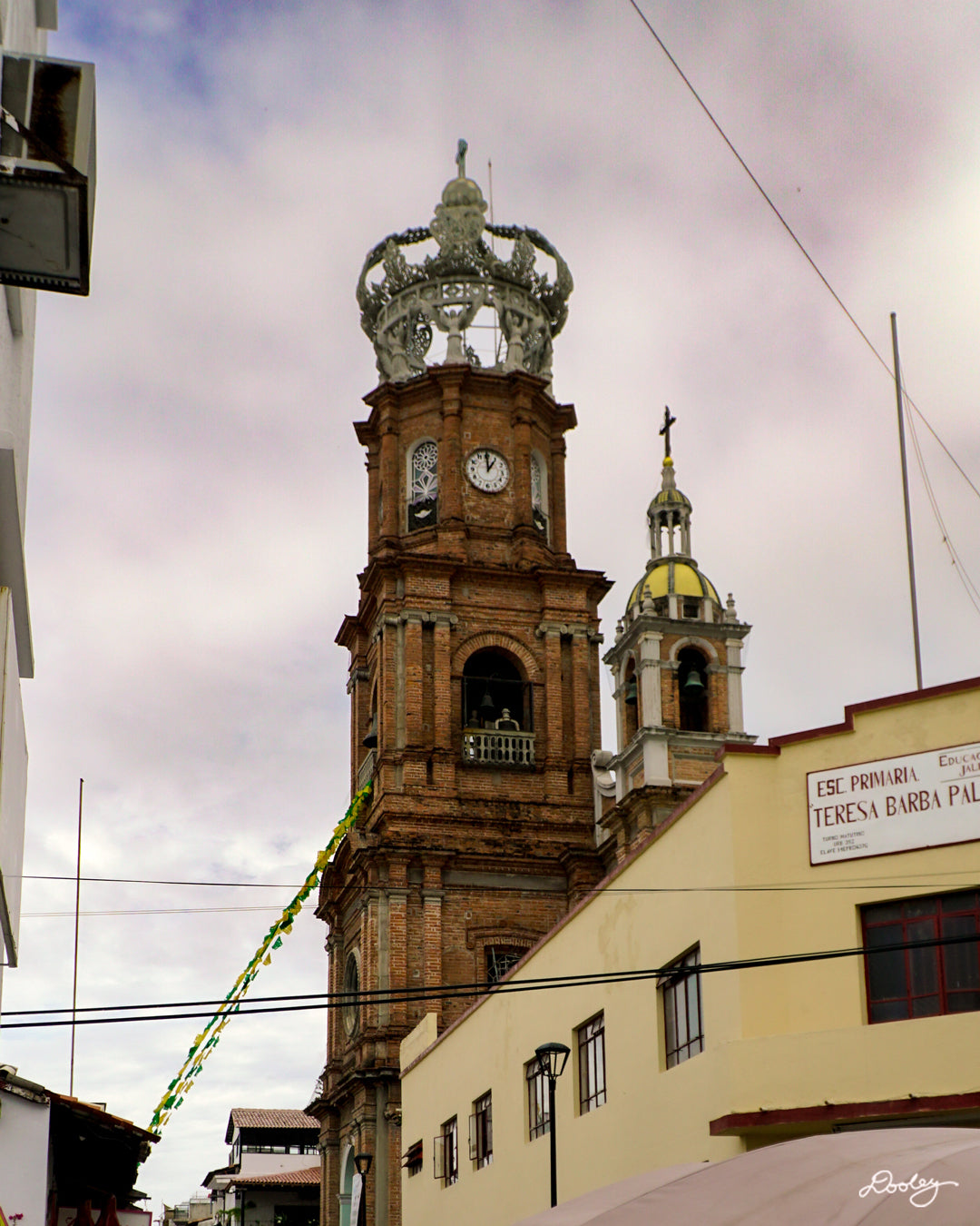 This striking photograph “Horchata Y Guadalupe” captures The Our Lady of Guadalupe Church from street level, showing it’s distinctive pink facade and its towering, ornate crown-shaped bell tower. The church stands tall against a cloudy blue sky, dominating the surrounding cityscape. The architectural details are intricate and beautiful, showcasing a blend of neoclassical and baroque styles. This print will add Puerto Vallarta’s culture to any space.