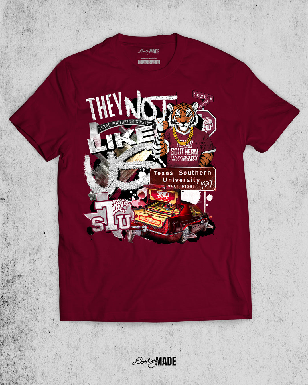A "They Not Like Us" themed Texas Southern University maroon shirt featuring a vibrant collage with a hand drawn and graphic elements: a tiger wearing a gold chain, Scott St and Wheeler Ave street sign, the "Texas Southern University Next Right" interstate sign, a spilled Styrofoam cup, a textured TSU logo, and a maroon cadillac with swangers and adapters. 