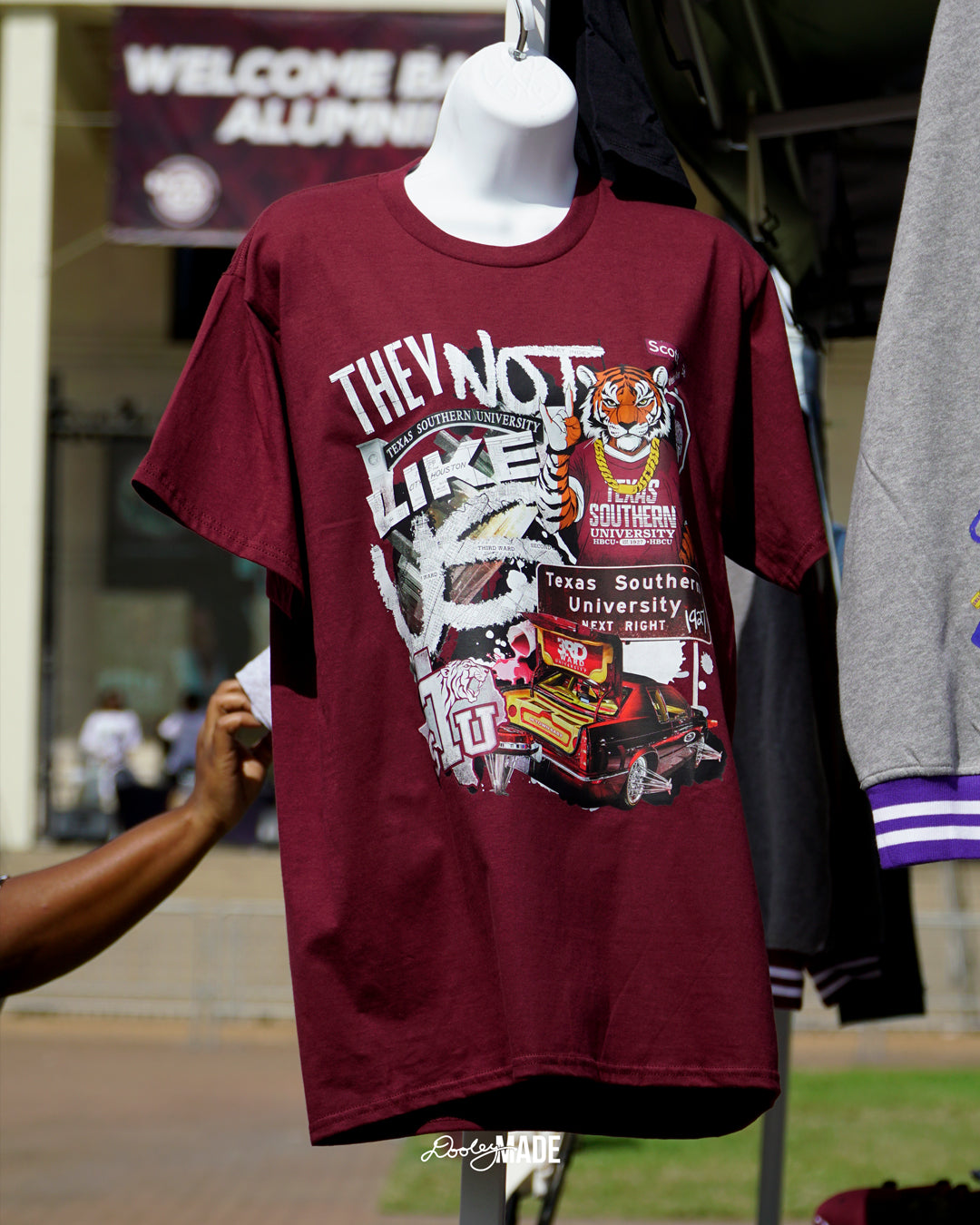 A photo of a A "They Not Like Us" themed Texas Southern University maroon shirt featuring a vibrant collage with a hand drawn and graphic elements: a tiger wearing a gold chain, Scott St and Wheeler Ave street sign, the "Texas Southern University Next Right" interstate sign, a spilled Styrofoam cup, a textured TSU logo, and a maroon cadillac with swangers and adapters. 