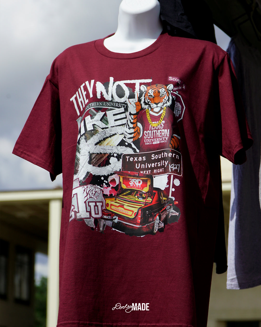 A photo of a A "They Not Like Us" themed Texas Southern University maroon shirt featuring a vibrant collage with a hand drawn and graphic elements: a tiger wearing a gold chain, Scott St and Wheeler Ave street sign, the "Texas Southern University Next Right" interstate sign, a spilled Styrofoam cup, a textured TSU logo, and a maroon cadillac with swangers and adapters. 