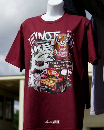 A photo of a A "They Not Like Us" themed Texas Southern University maroon shirt featuring a vibrant collage with a hand drawn and graphic elements: a tiger wearing a gold chain, Scott St and Wheeler Ave street sign, the "Texas Southern University Next Right" interstate sign, a spilled Styrofoam cup, a textured TSU logo, and a maroon cadillac with swangers and adapters. 