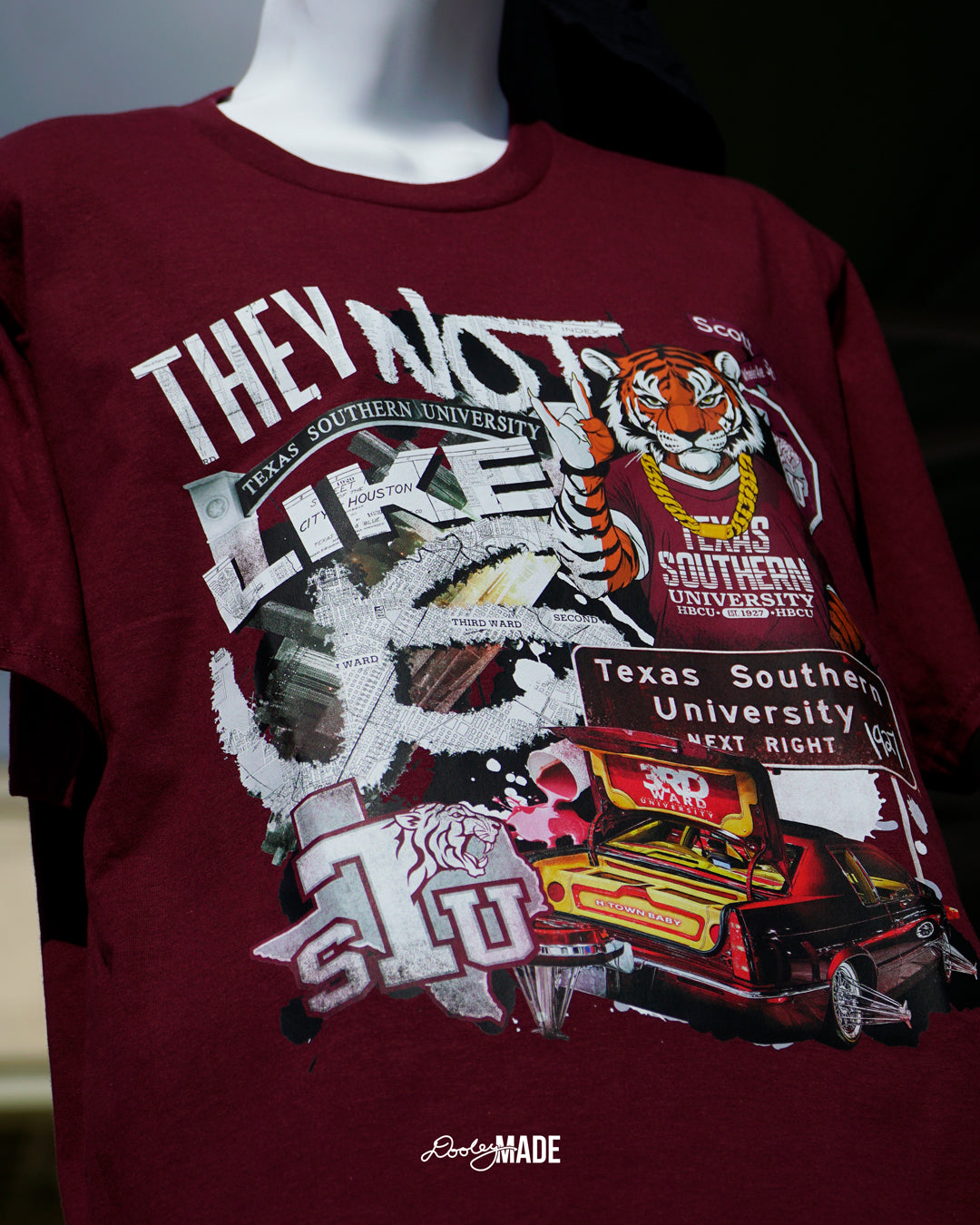 A photo of a A "They Not Like Us" themed Texas Southern University maroon shirt featuring a vibrant collage with a hand drawn and graphic elements: a tiger wearing a gold chain, Scott St and Wheeler Ave street sign, the "Texas Southern University Next Right" interstate sign, a spilled Styrofoam cup, a textured TSU logo, and a maroon cadillac with swangers and adapters. 