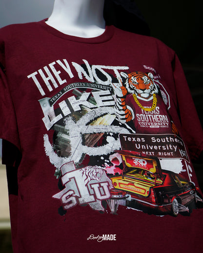 A photo of a A "They Not Like Us" themed Texas Southern University maroon shirt featuring a vibrant collage with a hand drawn and graphic elements: a tiger wearing a gold chain, Scott St and Wheeler Ave street sign, the "Texas Southern University Next Right" interstate sign, a spilled Styrofoam cup, a textured TSU logo, and a maroon cadillac with swangers and adapters. 