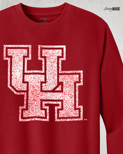 Image showing a red University of Houston crewneck sweatshirt with a white UH logo with red speckles stretched across the front, zoomed to show detail