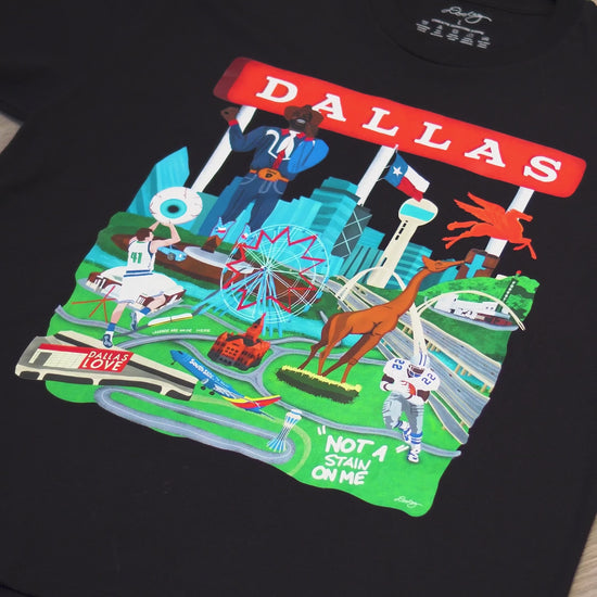 A video advertising a black Dallas themed shirt with the text "Dallas" in red and illustrated Dallas landmarks like the Dallas skyline, Big Tex and the Texas Star Ferris Wheel at the State Fair of Texas, legendary Dallas athletes, Dallas Love Field Airport, Dallas Zoo giraffe statue, and the Margaret Hunt Hill Bridge. Shirt is zoomed to show detail.