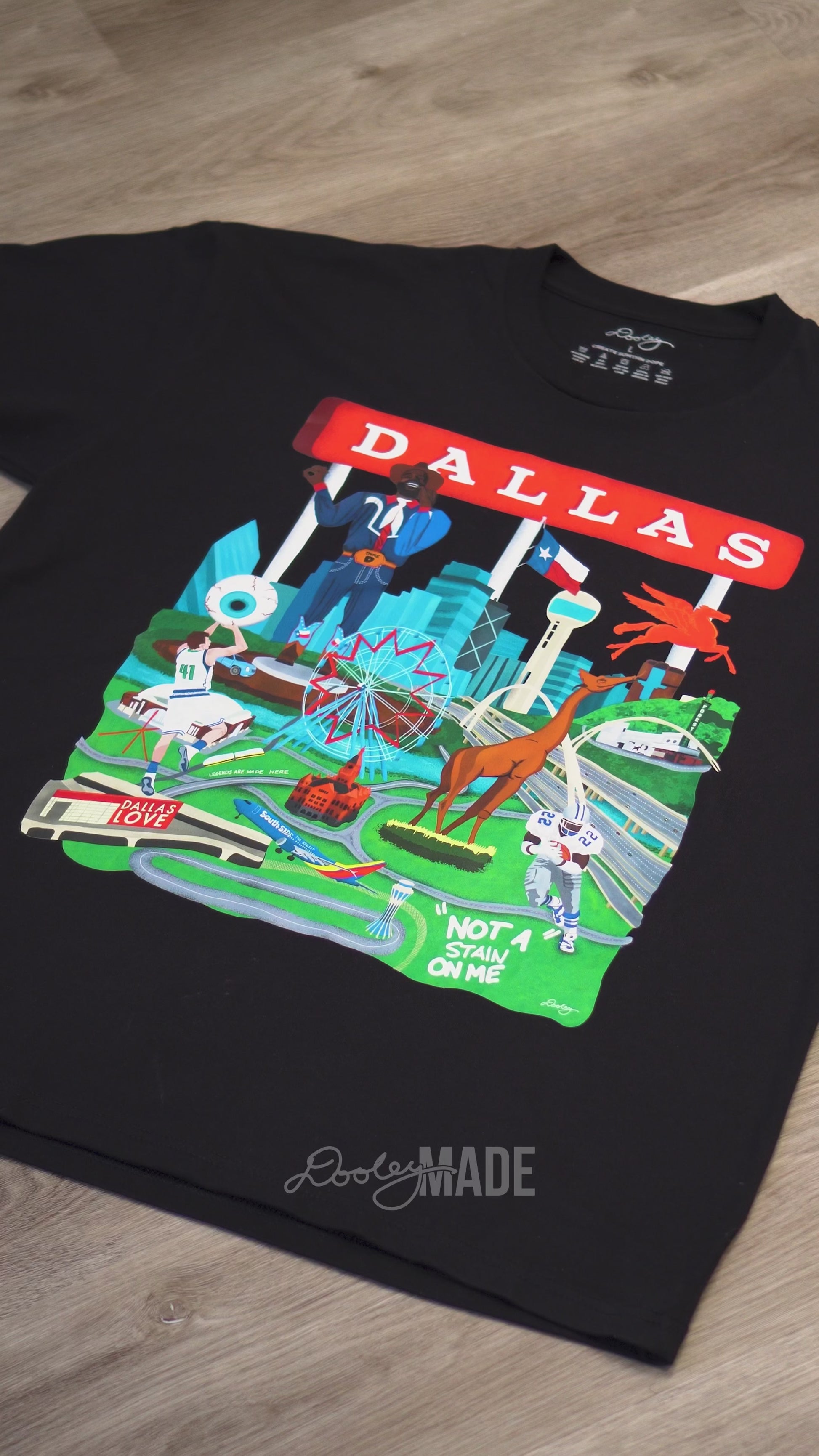 A video advertising a black Dallas themed shirt with the text "Dallas" in red and illustrated Dallas landmarks like the Dallas skyline, Big Tex and the Texas Star Ferris Wheel at the State Fair of Texas, legendary Dallas athletes, Dallas Love Field Airport, Dallas Zoo giraffe statue, and the Margaret Hunt Hill Bridge. Shirt is zoomed to show detail.