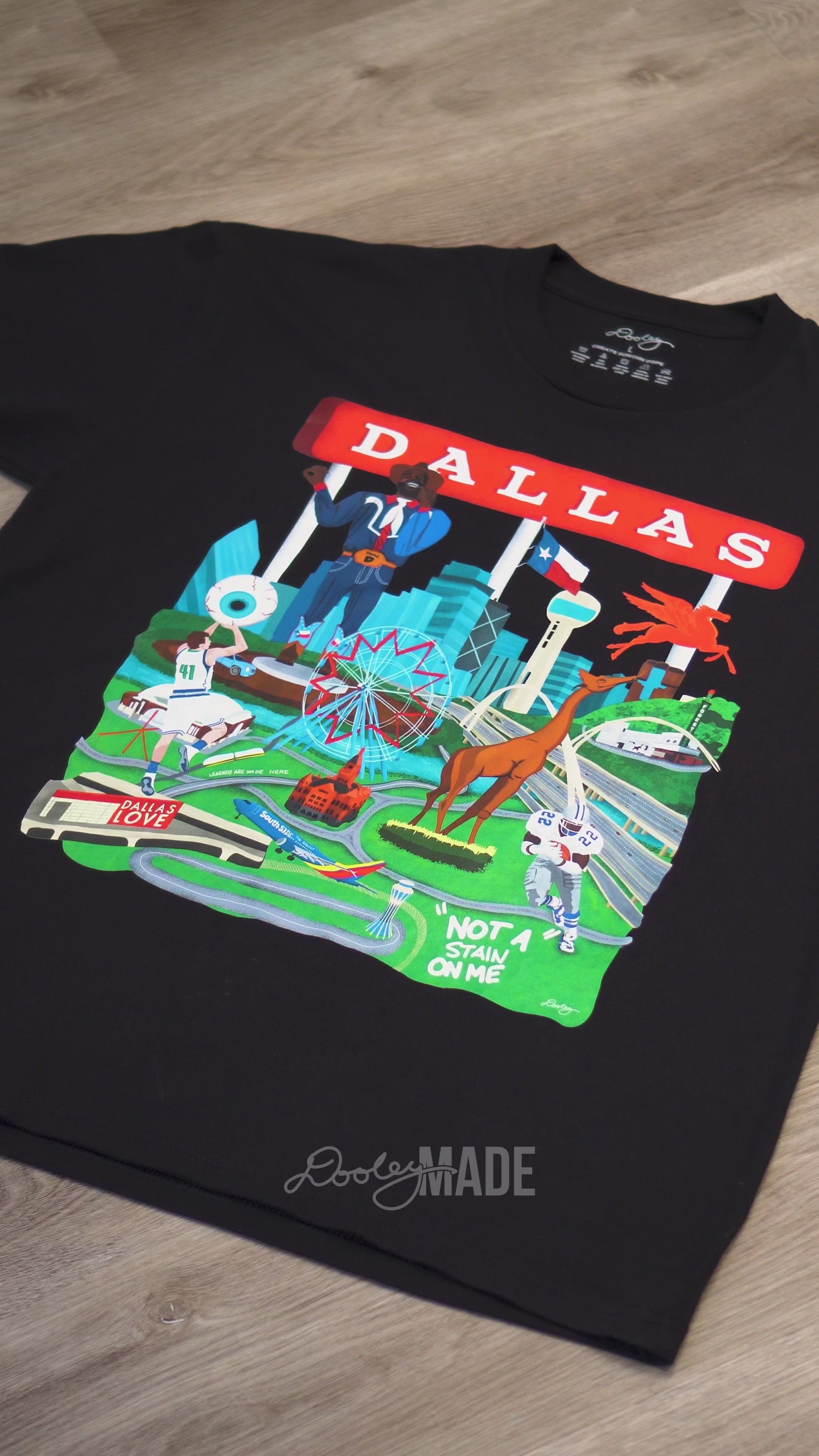 A video advertising a black Dallas themed shirt with the text "Dallas" in red and illustrated Dallas landmarks like the Dallas skyline, Big Tex and the Texas Star Ferris Wheel at the State Fair of Texas, legendary Dallas athletes, Dallas Love Field Airport, Dallas Zoo giraffe statue, and the Margaret Hunt Hill Bridge. Shirt is zoomed to show detail.